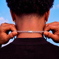 A close-up back view of a person wearing a silver chain necklace with a rectangular clasp, holding the chain in place with both hands. The person has curly black hair, gold hoop earrings, and is wearing a black ribbed shirt against a blue sky background.