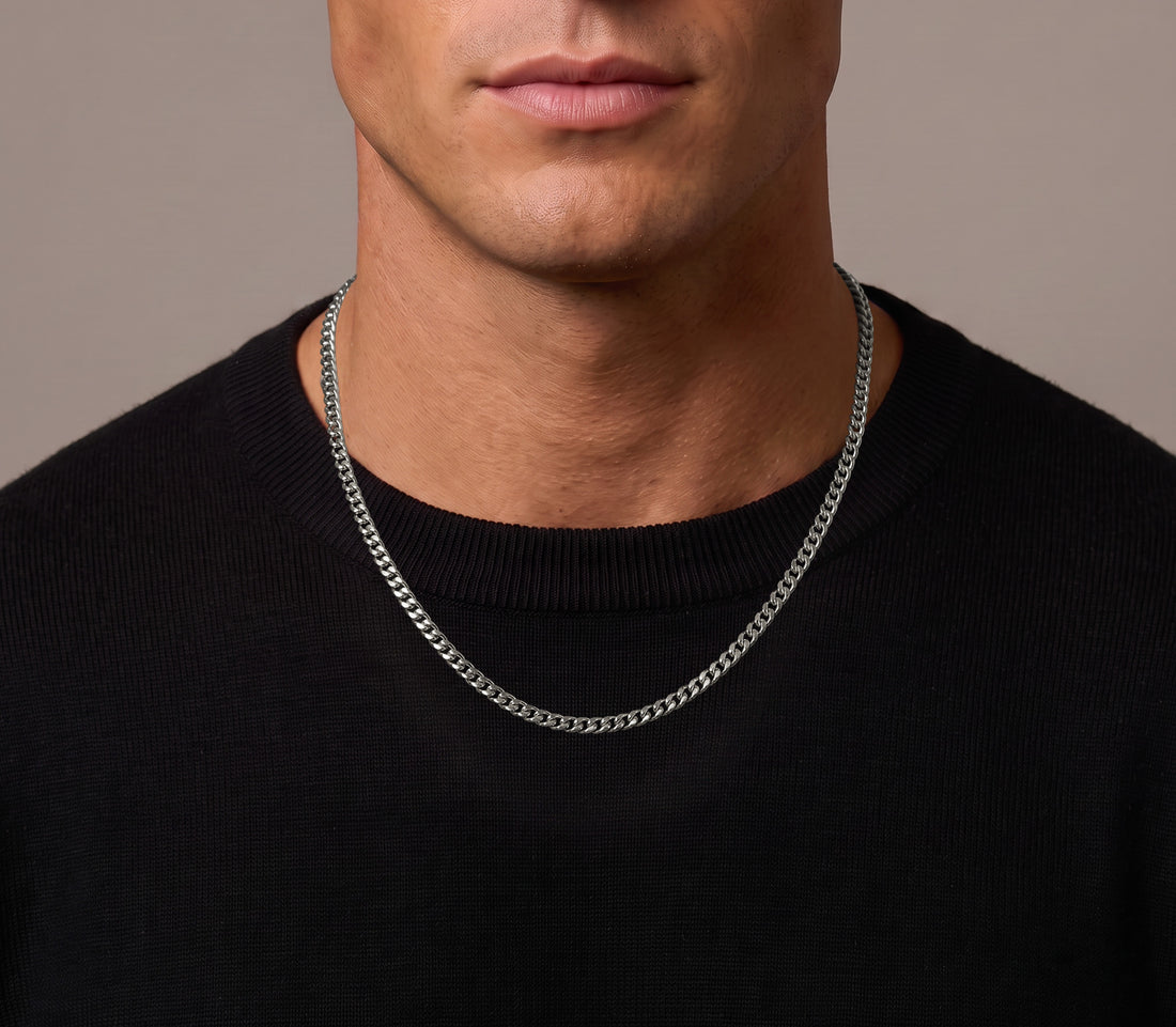 A close-up of a man wearing a silver chain necklace over a black crewneck sweater, set against a solid neutral background.